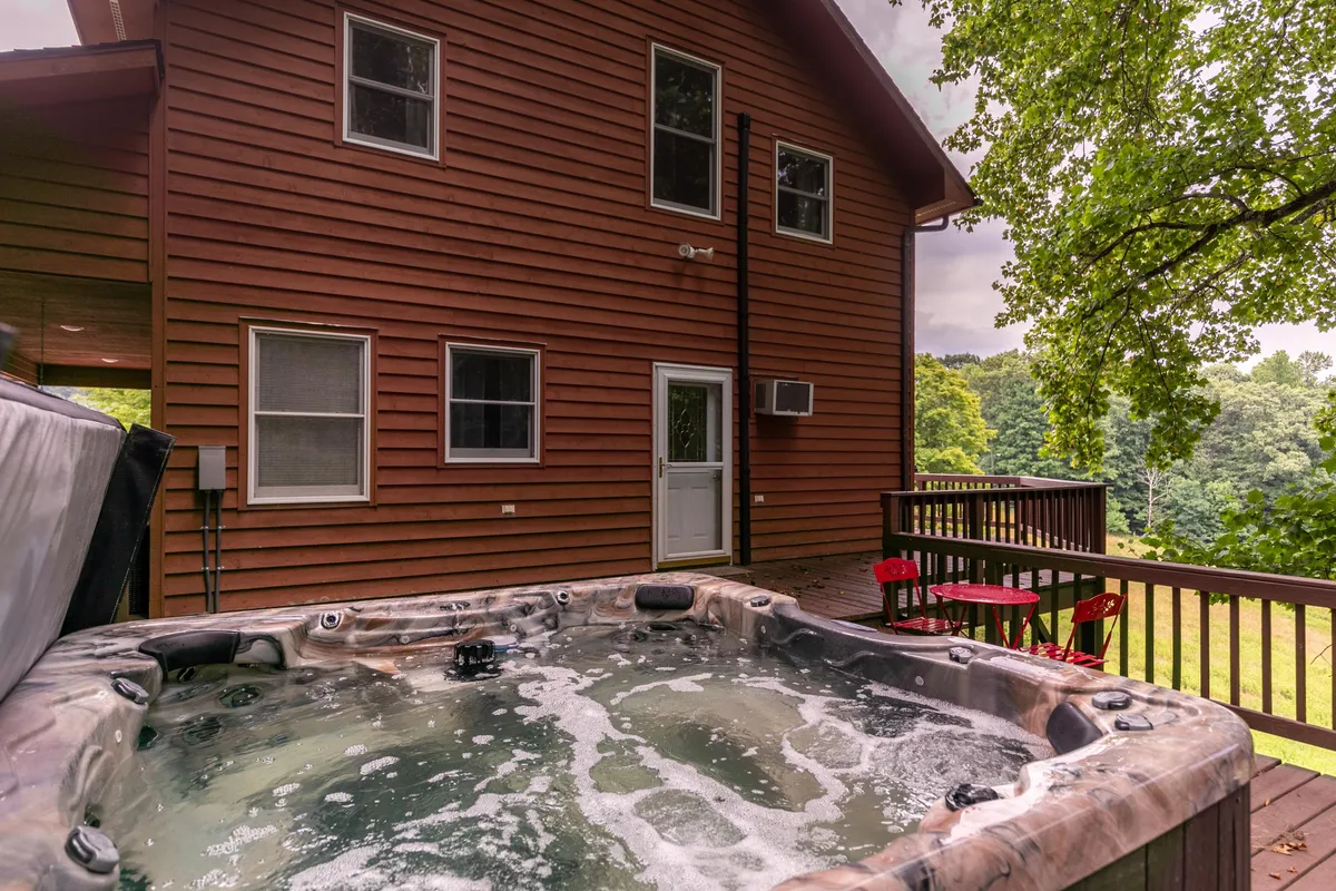 A wooden deck featuring a hot tub, red chairs, and the exterior of the home, with views of the surrounding landscape. The scene invites guests to unwind together, enjoy the views, or relax under the stars.