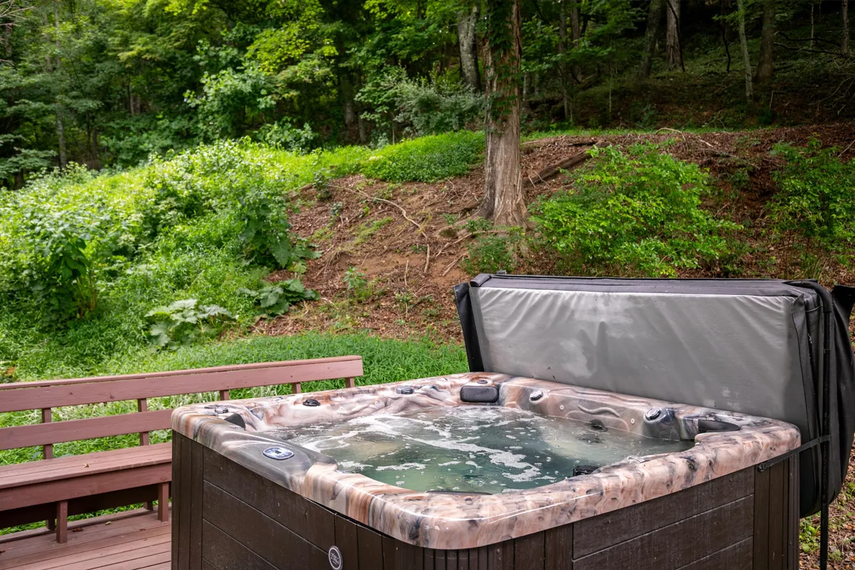The scene highlights an inviting hot tub and a wooden bench, surrounded by dense, lush greenery. The area radiates peace and relaxation, offering an ideal spot to unwind, stargaze, or enjoy the mountain air after outdoor excursions.