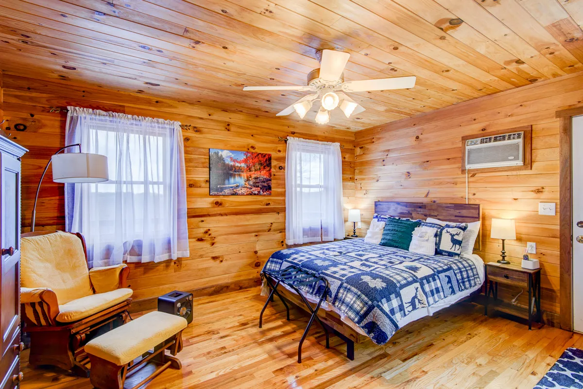 This image features a cozy bedroom with wooden walls, a bed adorned with a patterned blanket, an inviting armchair, and artwork on the wall. The setting is intimate and warm, communicating a sense of peaceful retreat ideal for rest and relaxation.