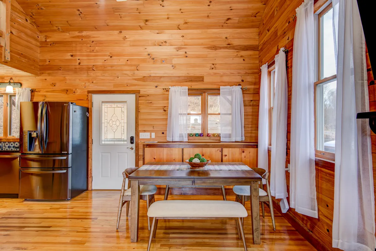 An open plan living and dining area with a wooden dining table set near large windows, modern kitchen appliances in the background, and natural wood paneling throughout. The space feels bright, cozy, and ready for memorable family meals.