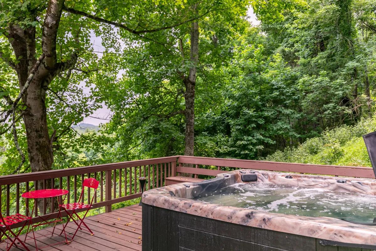 This image showcases a serene hot tub on a wooden deck, accompanied by red chairs and surrounded by lush greenery. The scene suggests a peaceful spot for relaxing, socializing, or soaking under the stars, with the beautiful backdrop of nature all around.