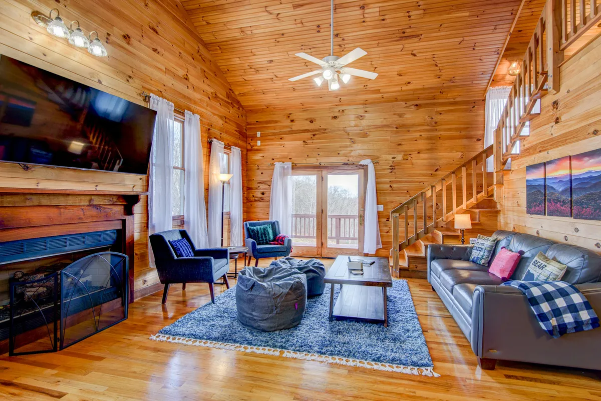 A cozy living room with a large plush sofa adorned with throw pillows, a bean bag chair, a glowing gas fireplace beneath a wooden ceiling, large windows with curtains, and a prominent flat-screen TV. The space is arranged for relaxation and socializing.