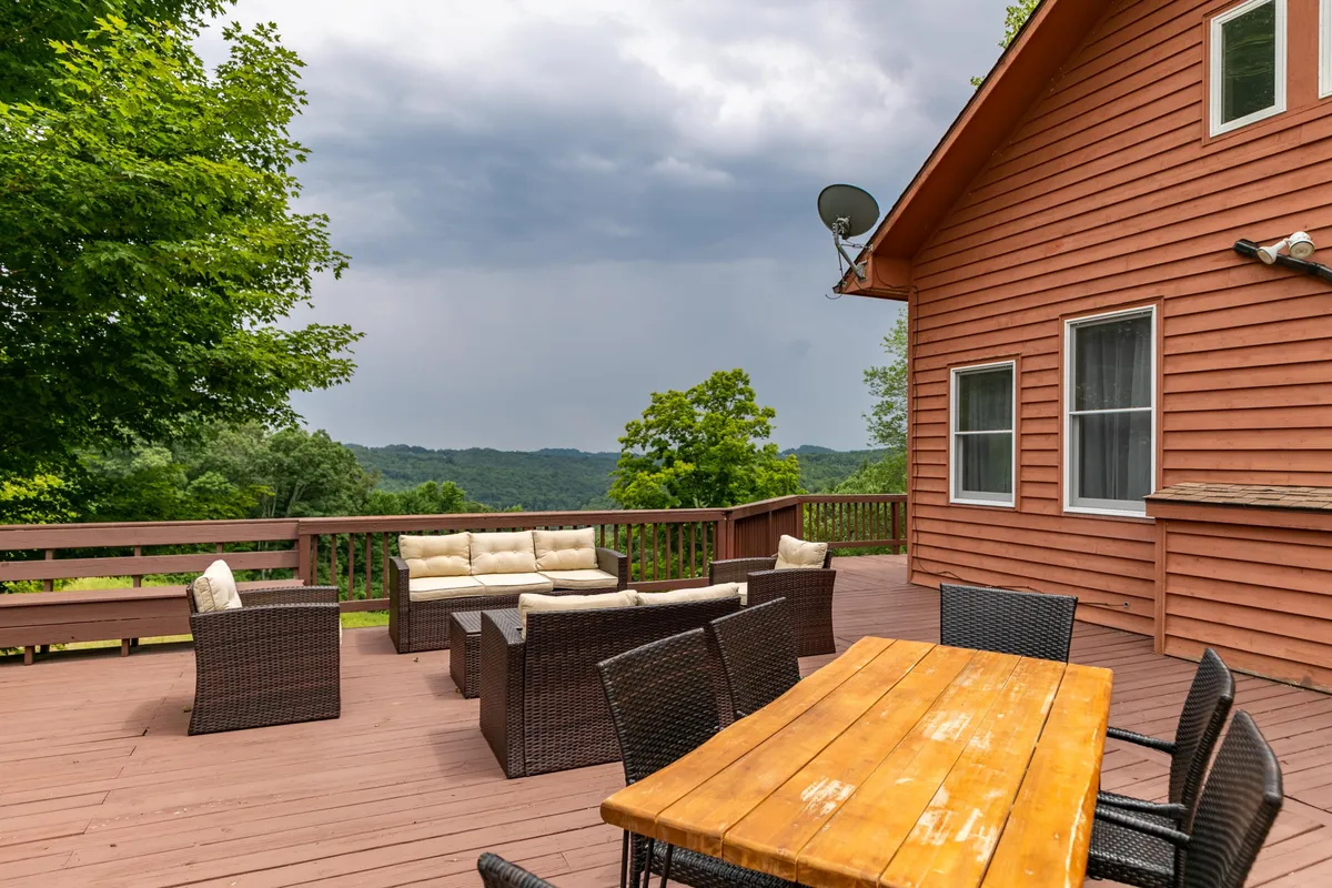 A spacious outdoor seating area with a sturdy wooden table set for dining, surrounded by comfortable chairs. Rolling hills and lush greenery create a stunning backdrop, and the area is positioned to capture beautiful sunset views. The setting invites families to gather for meals, games, or relaxed conversation while enjoying the natural mountain landscape.