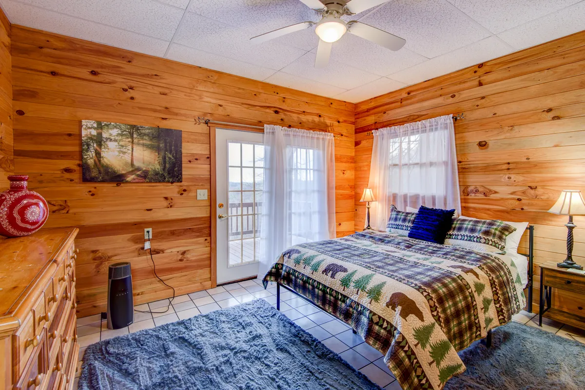 The image showcases a bedroom with a patterned quilt, large windows letting in natural light, wooden walls, a dresser with a decorative vase, and a nightstand with a lamp. The setting is serene and bright, suggesting restful mornings and cozy evenings.