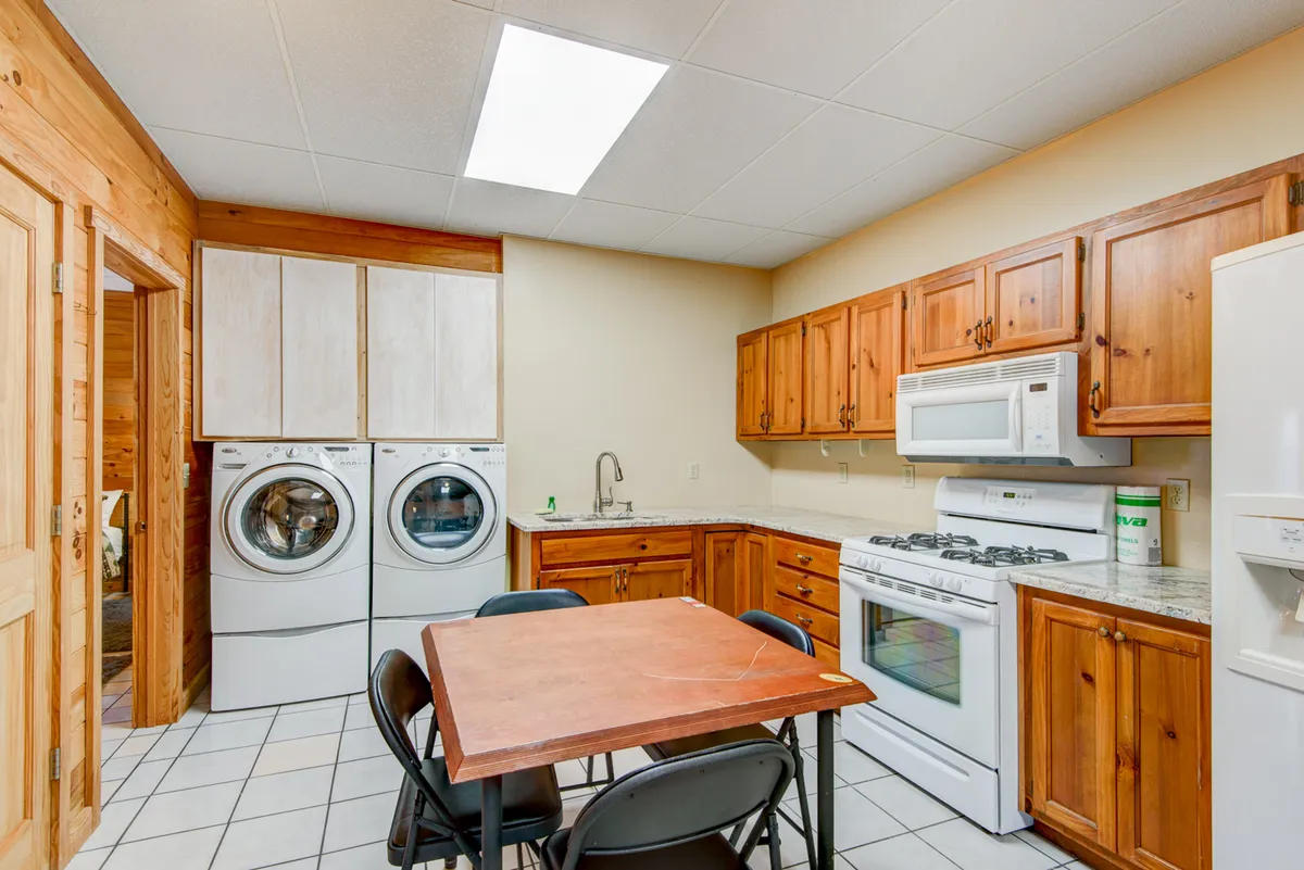 This interior image shows a practical kitchen-dining area with a visible washing machine, a kitchen table, wooden cabinets, and kitchen appliances. The space feels homey and functional, clearly illustrating the combined utility of kitchen and laundry in one area.